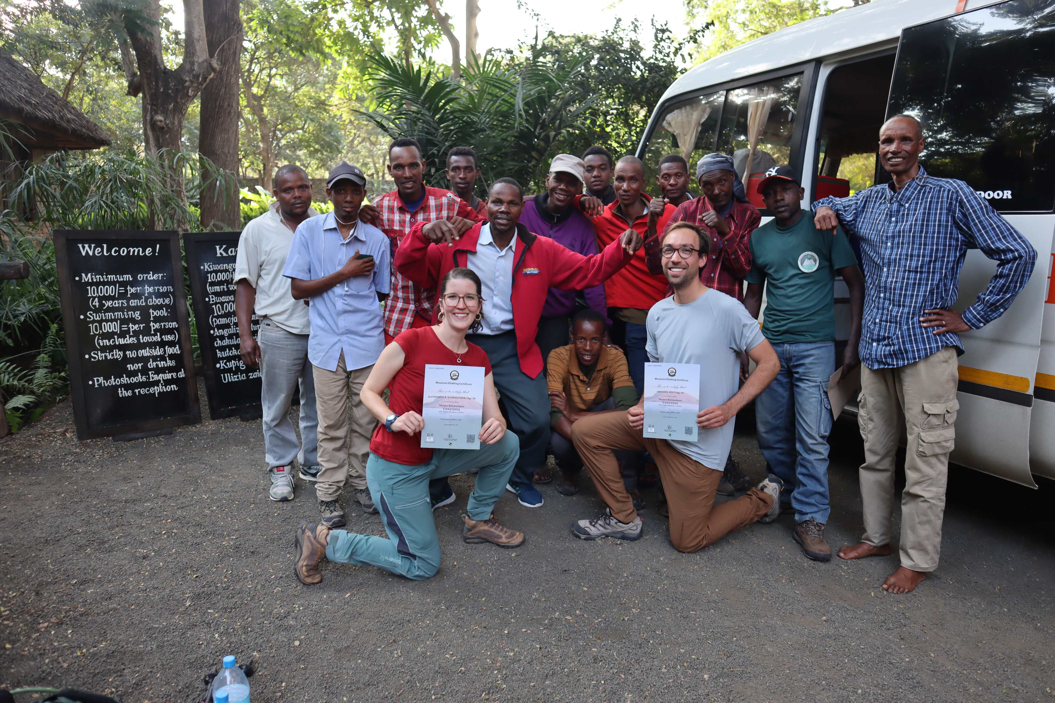 Katherine and Tim showing off their certificates with the team in front of a bus at the entrance to Rivertrees