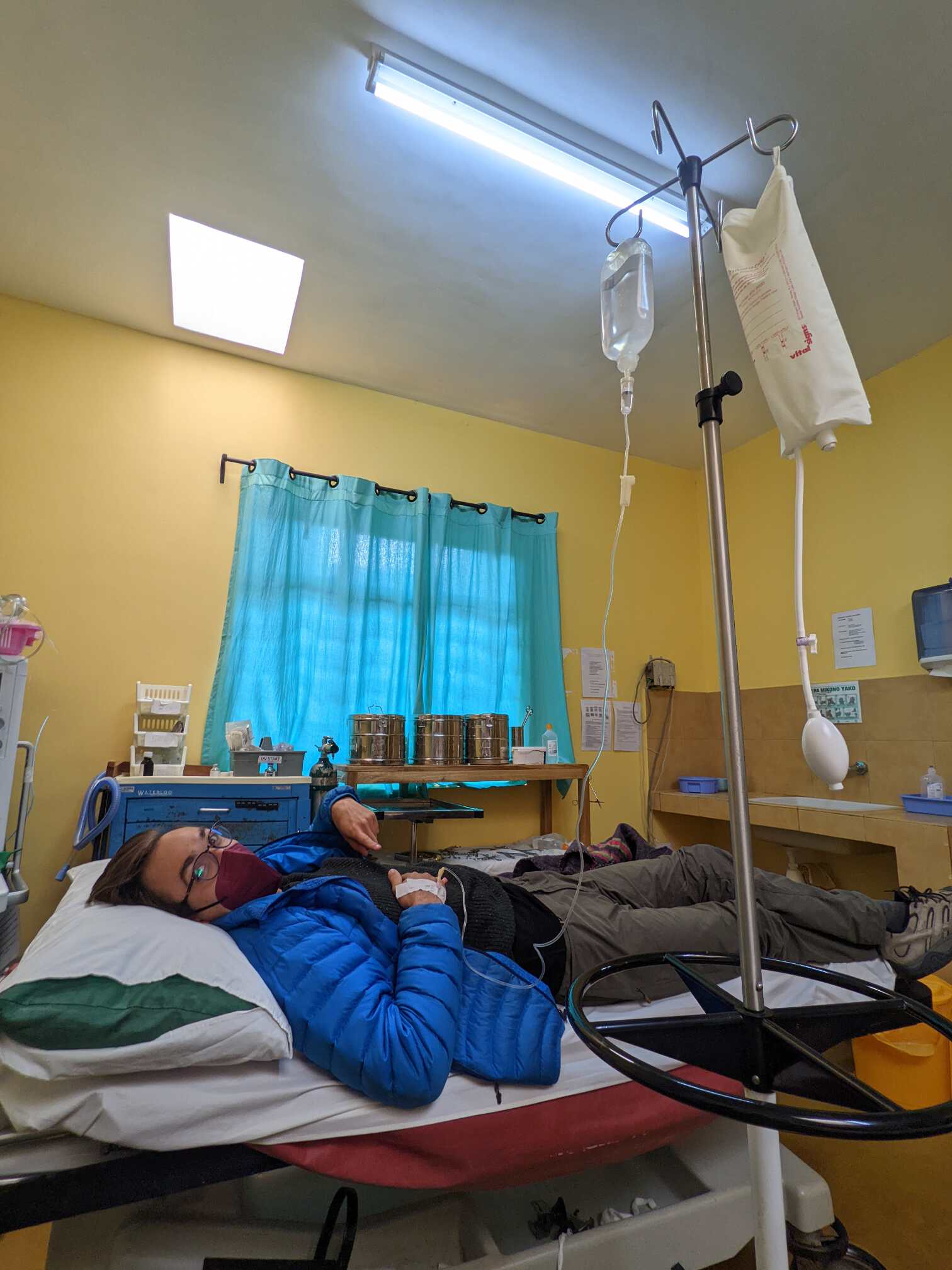 Tim wearing a winter coat in a hospital bed pointing to an IV drip in his right hand