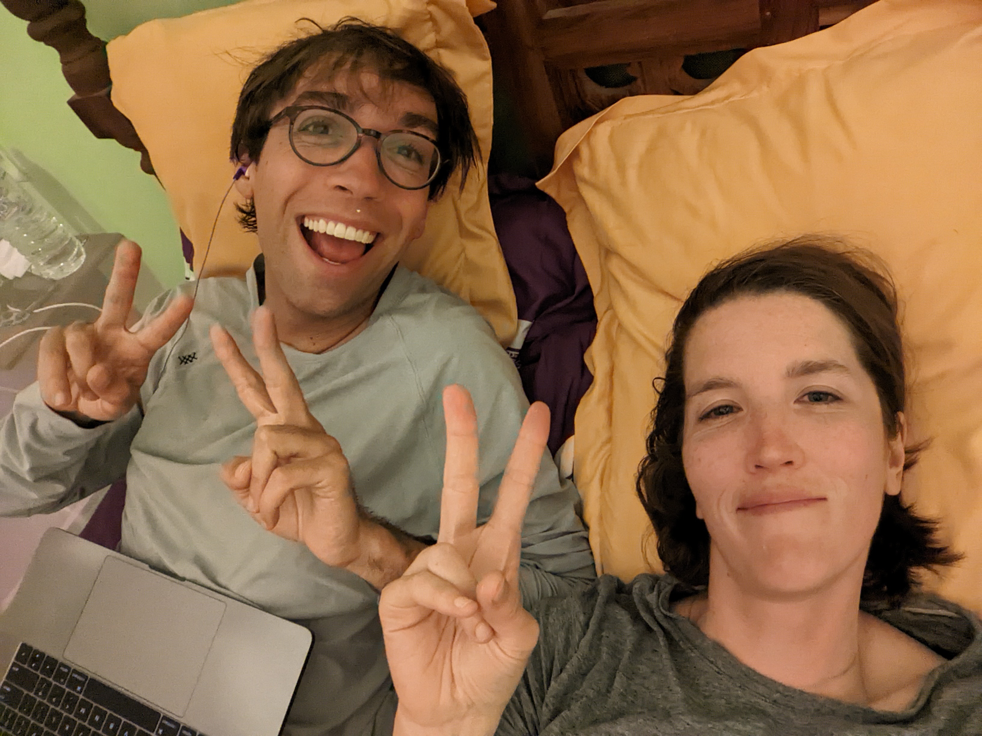 Kat and Tim give peace signs while lying in bed watching TV on their computer